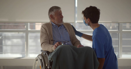 Nurse in safety mask interacting with disabled senior patient in wheelchair in corridor at hospital.