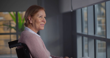Mature woman sitting in wheelchair in nursing home looking out of windowの写真素材
