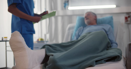 Doctor talking to senior man patient with broken leg lying in hospital bedの写真素材