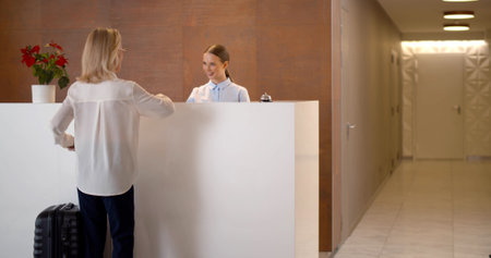 Senior woman with luggage taking room key card from female staff at reception desk in hotelの写真素材