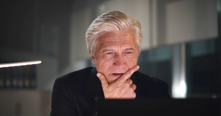 Professional mature businessman sitting in dark office and working on laptopの写真素材