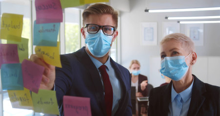 Business colleagues in protective face mask writing on sticky note on glass wallの写真素材