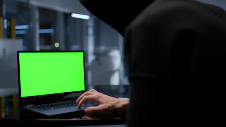 Close up of hacker in hoodie working on laptop with green screen in dark officeの写真素材