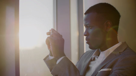 Young black businessman taking picture of sunset on smartphone standing near window in officeの写真素材