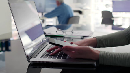 Close up of businesswoman holding smartphone and typing on laptop at workplace