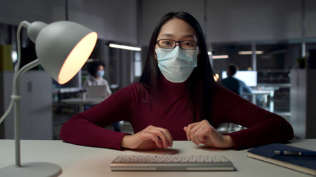 Female employee in safety mask sit at desk typing on computer keyboardの写真素材