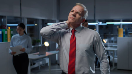 Mature businessman suffer from pain in neck standing late in officeの写真素材