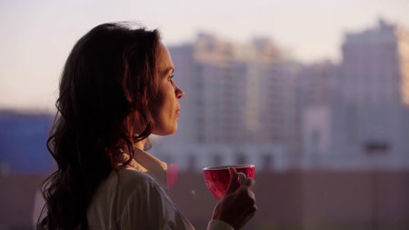 Young beautiful businesswoman drink coffee looking ut of window at sunset in modern officeの写真素材