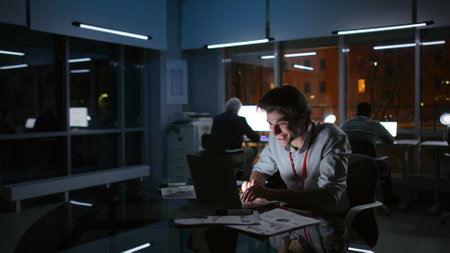 Portrait of happy businessman looking at laptop and laughing working late at night in modern officeの写真素材
