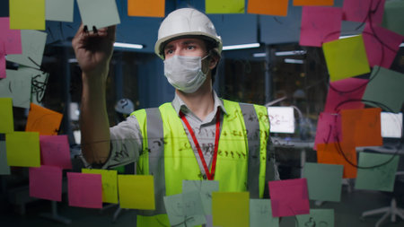Engineer in hardhat and safety mask write on glass board with sticky notes in officeの写真素材