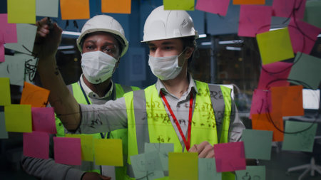 Diverse architects in safety mask and hardhat discuss and write on glass board with sticky notesの写真素材