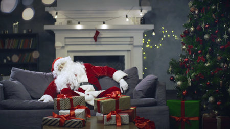 Tired Santa Claus sleeping on couch after packing christmas presents in living roomの写真素材