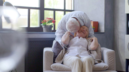 Smiling aged woman sitting in armchair and husband kissing herの写真素材