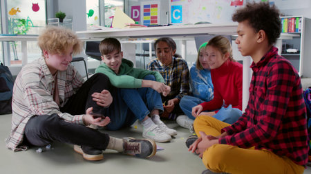 Multiethnic teen classmates sit on floor in class and watch video on smartphone together