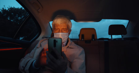 Businessman wearing face mask sitting on backseat of car and using smartphoneの写真素材