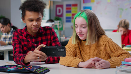 Diverse teenage classmates watch video on smartphone sitting at desk in classroomの写真素材