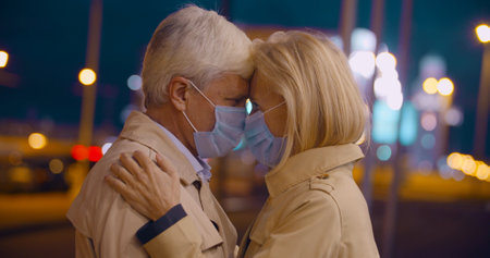 Bokeh shot of senior couple in face mask hugging on city street at nightの写真素材