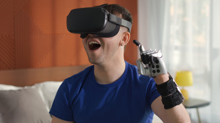Young man contemporary bionic arm use virtual reality headset playing video game sitting on bedの写真素材