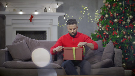 Portrait of man packing Christmas present sitting on couch in decorated living room.の写真素材