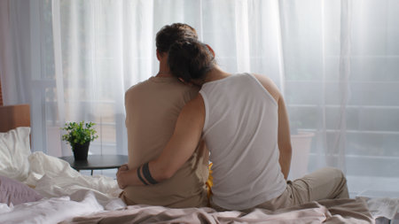 Back view of diverse homosexual couple sitting on bed and huggingの写真素材