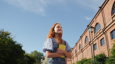 Low angle view of smiling teenage girl with book and backpack standing outside school buildingの写真素材