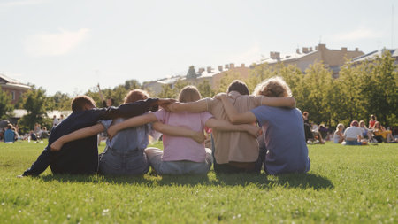 Back view of diverse teen students sit on school lawn and hugの写真素材