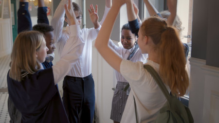 Diverse teenagers making stack of hands and hugging in school corridor.