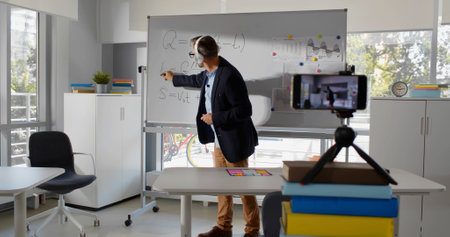 Mature physics teacher in headphones writing on whiteboard making tutorial video on smartphoneの写真素材