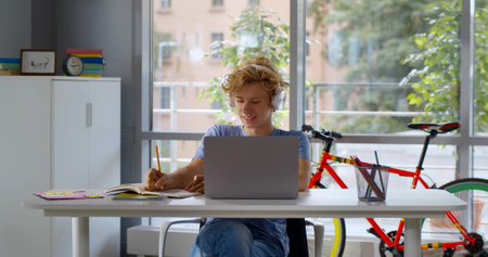 Portrait of teen boy in headphones doing homework on laptop at home. Caucasian teenage student watching online webinarの写真素材