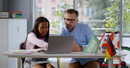 Mature teacher and african schoolgirl study foreign language on laptop in classroomの写真素材