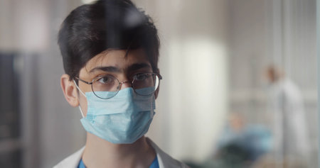 Young doctor in protective mask looking out of window in hospital