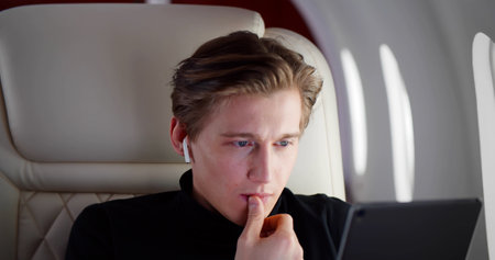 Young handsome billionaire in earphones using digital tablet flying first class