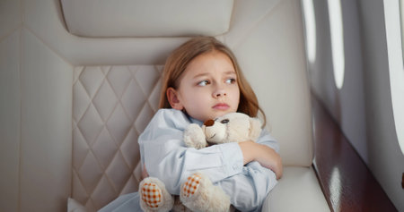 Scared little girl hugging teddy bear sitting in leather chair of business class airplane cabin