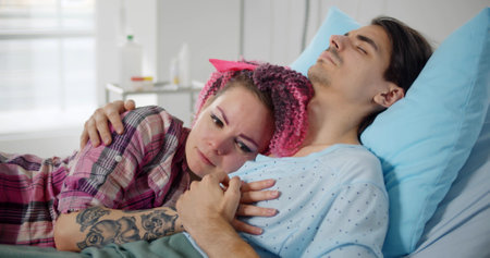 Worried and sad young woman hugging sick husband sleeping in hospital bedの写真素材