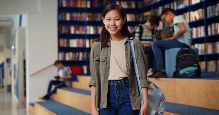 Portrait of smiling asian schoolgirl looking at camera standing in libraryの写真素材