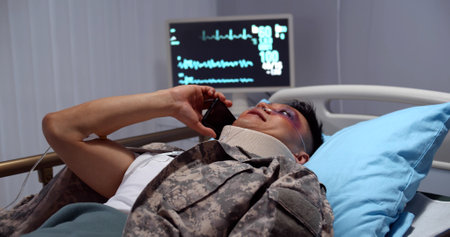 Injured asian soldier with neck collar lying in hospital bed and talking on smartphoneの写真素材