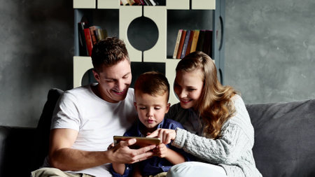 Young happy couple sitting with child oon sofa using tablet at home