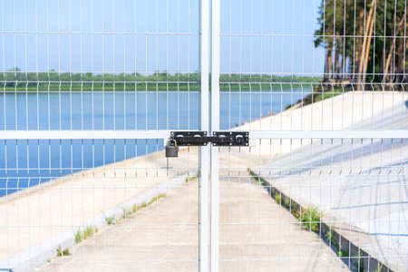 The fence is vertical metal (gate) on the background of the river, the sky, closed with a padlock. A territory free of COVID-19. Concept: private property, fenced territory. No entry allowedの写真素材