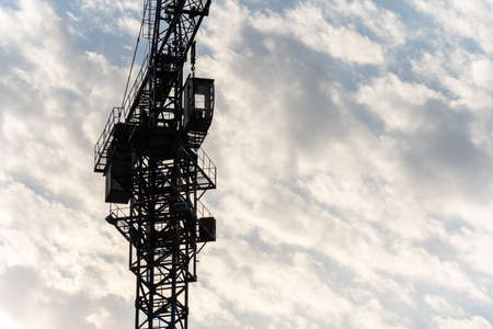 A large fragment of a tower crane with a cabin and an arrow against the background of clouds and blue sky. Machine for high-altitude lifting of building materialsの写真素材