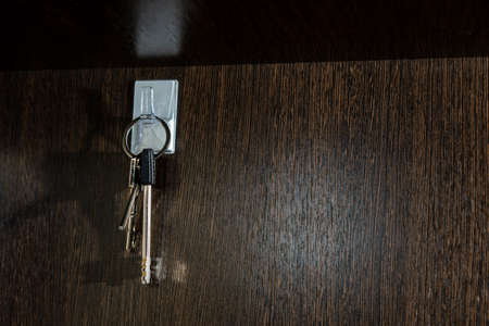 A bunch of three metal keys, fastened with a ring, hangs on a light hook on a smooth, even, dark, wooden surface, casting a shadowの写真素材