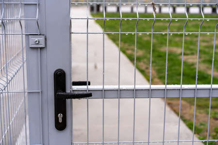 Black lock with a handle in a lattice gate made of metal rods of gray color with raindrops, behind which you can see an asphalt path and grass on an autumn day. Blur the far backgroundの写真素材