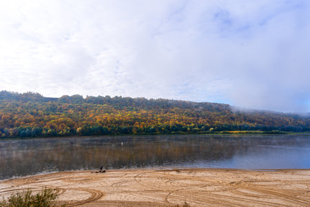 A view of the river between two banks, a fisherman on the shore and in a boat, a colorful forest on...