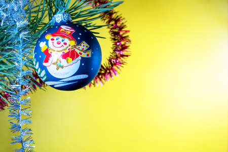 Christmas tree glass toy in the form of a blue glass ball with a picture of a smiling snowman in a hat hanging under a green fir branch with tinsel on a yellow backgroundの写真素材