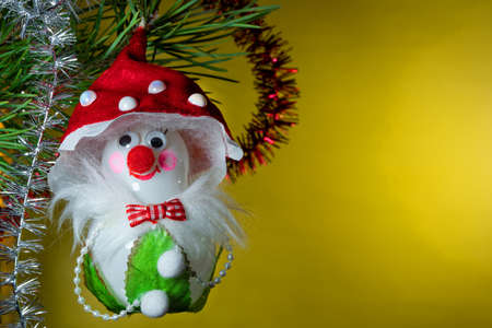 Christmas tree glass toy in the form of a smiling mushroom with a red nose, eyes and a red hat, hanging under a fir branch on a background with tinsel on a yellow backgroundの写真素材