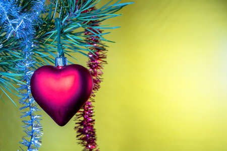 Christmas tree glass toy in the form of a red heart hanging under a spruce green branch with tinsel on a yellow backgroundの写真素材