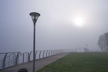 The morning sun breaks through the fog. A landscape with a fenced-in metal fence, a street lamp, an urn and green grass. Nature backgroundの写真素材