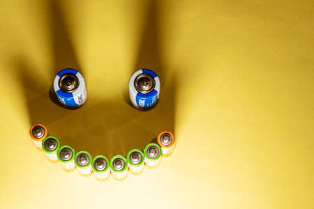 Top view of a face made of used electric cylindrical batteries and shadows on a bright yellow background. Free space for copying, design or text. The concept of ecology, waste disposal and recyclingの写真素材