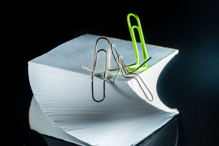 A view of two curved metal paper clips sitting on a twisted paper block for notes and hugging. A green paper clip came up to them. The background is black. The concept of communicationの写真素材