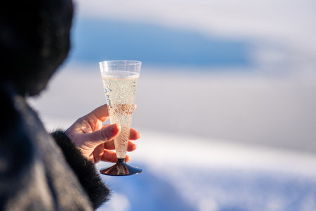 A view of a woman's hand, dressed in a mink fur coat, holding a glass of wine on a sunny winter day. The background is blurred. Free space for design, text, copyingの写真素材