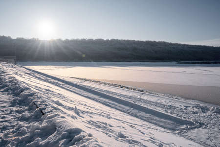River landscape with a view of the sun on a December winter day over a hill covered with forest and a frozen riverの写真素材